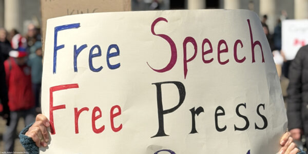 Person at a rally holds a large handmade sign reading “Free Speech, Free Press, Free People,” with a crowd and the Lincoln Memorial building in the background.