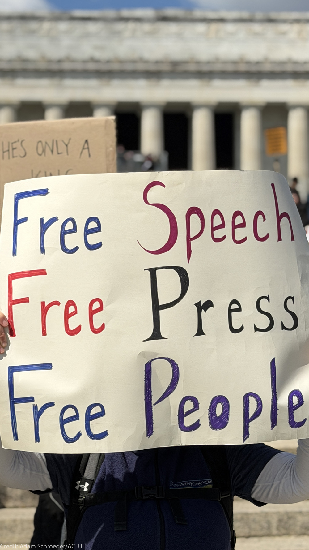 Person at a rally holds a large handmade sign reading “Free Speech, Free Press, Free People,” with a crowd and the Lincoln Memorial building in the background.