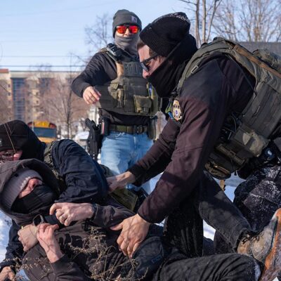 Federal immigration agents in tactical gear tackle a protester to the ground in the snow beside a chain-link fence, while other agents stand nearby in a residential area with houses and a school bus in the background.
