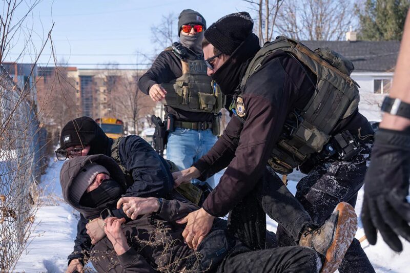 Federal immigration agents in tactical gear tackle a protester to the ground in the snow beside a chain-link fence, while other agents stand nearby in a residential area with houses and a school bus in the background.