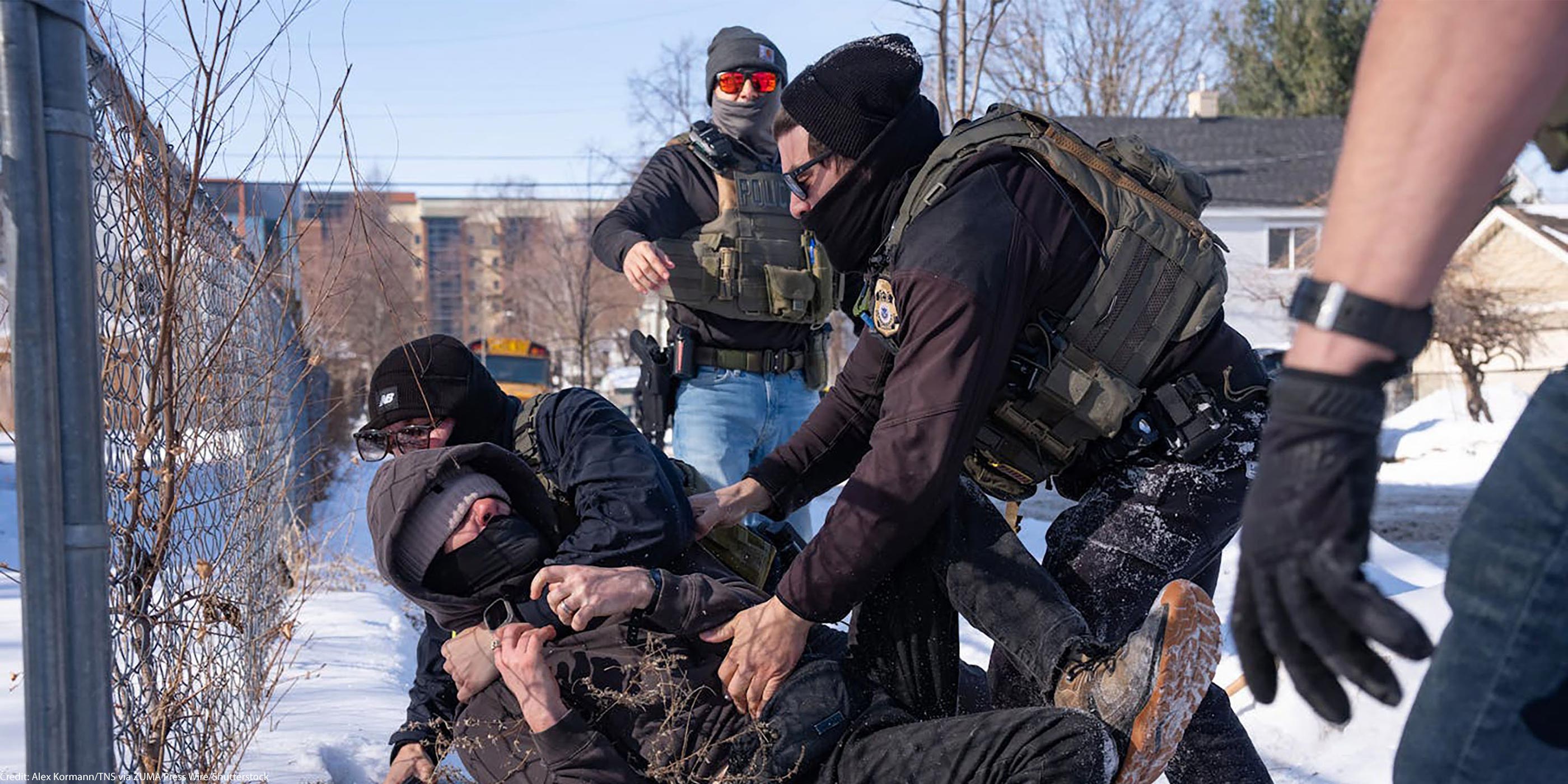 Federal immigration agents in tactical gear tackle a protester to the ground in the snow beside a chain-link fence, while other agents stand nearby in a residential area with houses and a school bus in the background.
