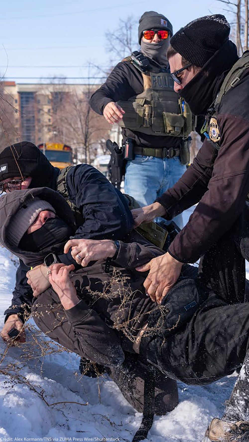Federal immigration agents in tactical gear tackle a protester to the ground in the snow beside a chain-link fence, while other agents stand nearby in a residential area with houses and a school bus in the background.