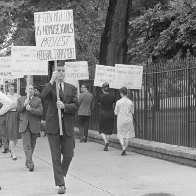 Black-and-white photo of an LGBTQ rights picket on a tree-lined sidewalk beside a tall iron fence (near the White House). Protesters—mostly in suits and dresses—carry hand-lettered signs including “FIFTEEN MILLION U.S. HOMOSEXUALS PROTEST FEDERAL TREATMENT,” “FIRST-CLASS CITIZENSHIP FOR HOMOSEXUALS,” and “SEXUAL PREFERENCE IS IRRELEVANT TO EMPLOYMENT.” A uniformed police officer stands with hands on hips facing the group, while an ABC TV reporter in a suit holds a microphone at the curb; pedestrians walk past in the background.