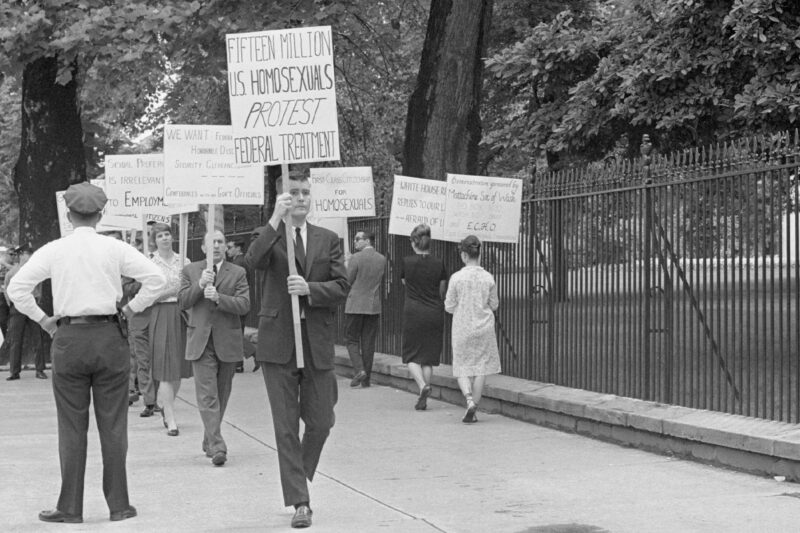 Black-and-white photo of an LGBTQ rights picket on a tree-lined sidewalk beside a tall iron fence (near the White House). Protesters—mostly in suits and dresses—carry hand-lettered signs including “FIFTEEN MILLION U.S. HOMOSEXUALS PROTEST FEDERAL TREATMENT,” “FIRST-CLASS CITIZENSHIP FOR HOMOSEXUALS,” and “SEXUAL PREFERENCE IS IRRELEVANT TO EMPLOYMENT.” A uniformed police officer stands with hands on hips facing the group, while an ABC TV reporter in a suit holds a microphone at the curb; pedestrians walk past in the background.