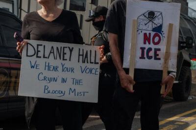 People hold signs outside the Delaney Hall Detention Facility during a protest against the treatment of detainees; one sign reads "Delaney Hall, we hear your voice cryin’ in the bloody mist," and another says "NO ICE" beneath a skull-and-crossbones drawing, with a masked officer and parked vehicles behind them.