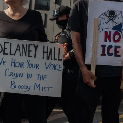 People hold signs outside the Delaney Hall Detention Facility during a protest against the treatment of detainees; one sign reads "Delaney Hall, we hear your voice cryin’ in the bloody mist," and another says "NO ICE" beneath a skull-and-crossbones drawing, with a masked officer and parked vehicles behind them.