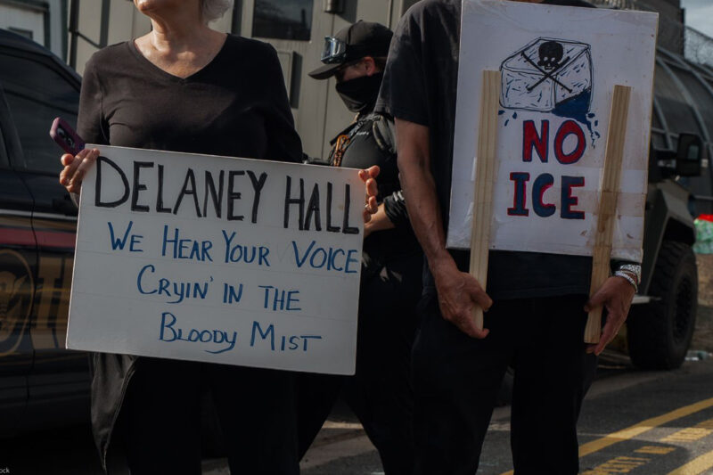 People hold signs outside the Delaney Hall Detention Facility during a protest against the treatment of detainees; one sign reads "Delaney Hall, we hear your voice cryin’ in the bloody mist," and another says "NO ICE" beneath a skull-and-crossbones drawing, with a masked officer and parked vehicles behind them.