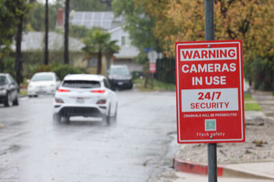 Rain falls on a residential street, where a red security sign warns "Cameras in Use 24/7 Security, Criminals Will Be Prosecuted, and flock safety." Cars drive down the wet road past houses, trees, sidewalks, and fences in the background.