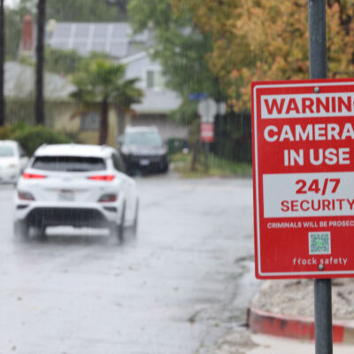 Rain falls on a residential street, where a red security sign warns "Cameras in Use 24/7 Security, Criminals Will Be Prosecuted, and flock safety." Cars drive down the wet road past houses, trees, sidewalks, and fences in the background.