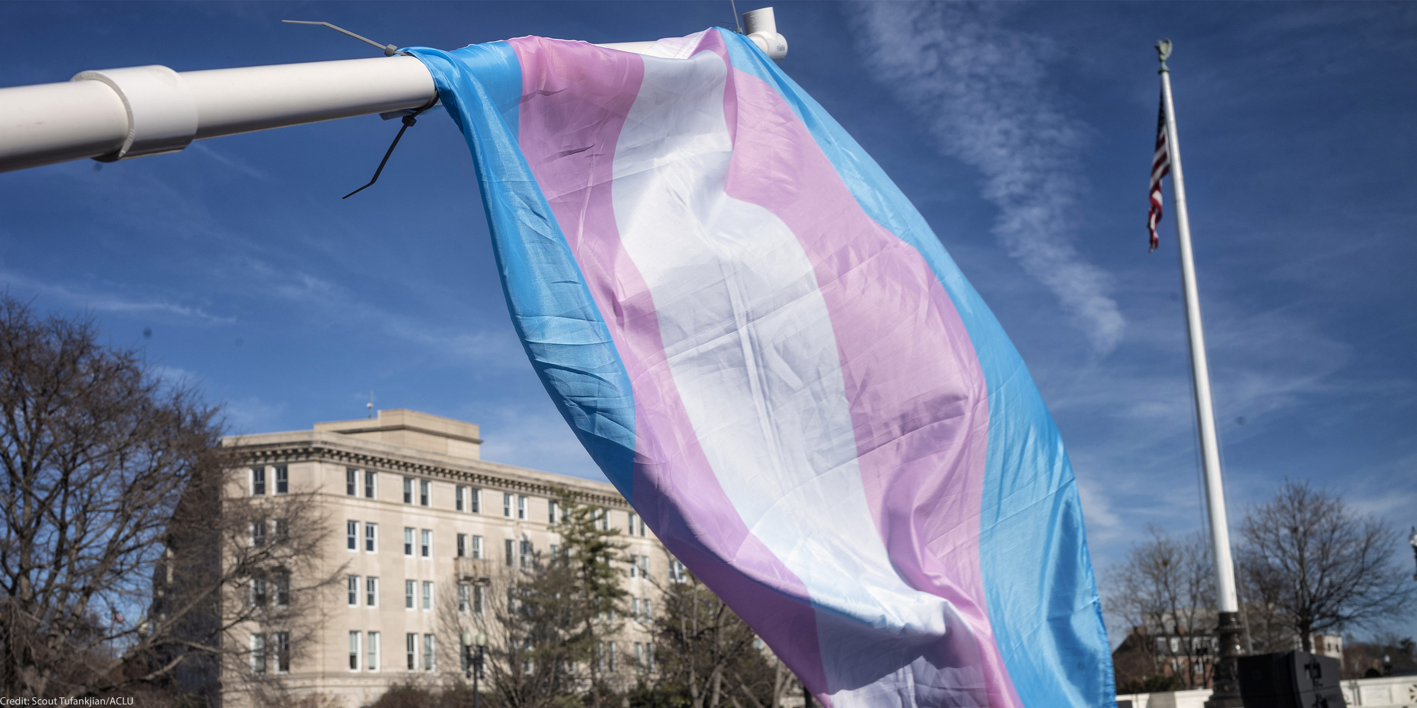 A large transgender pride flag waves on a flagpole against a blue sky, with an American flag flying on a tall pole and a government building in the background.
