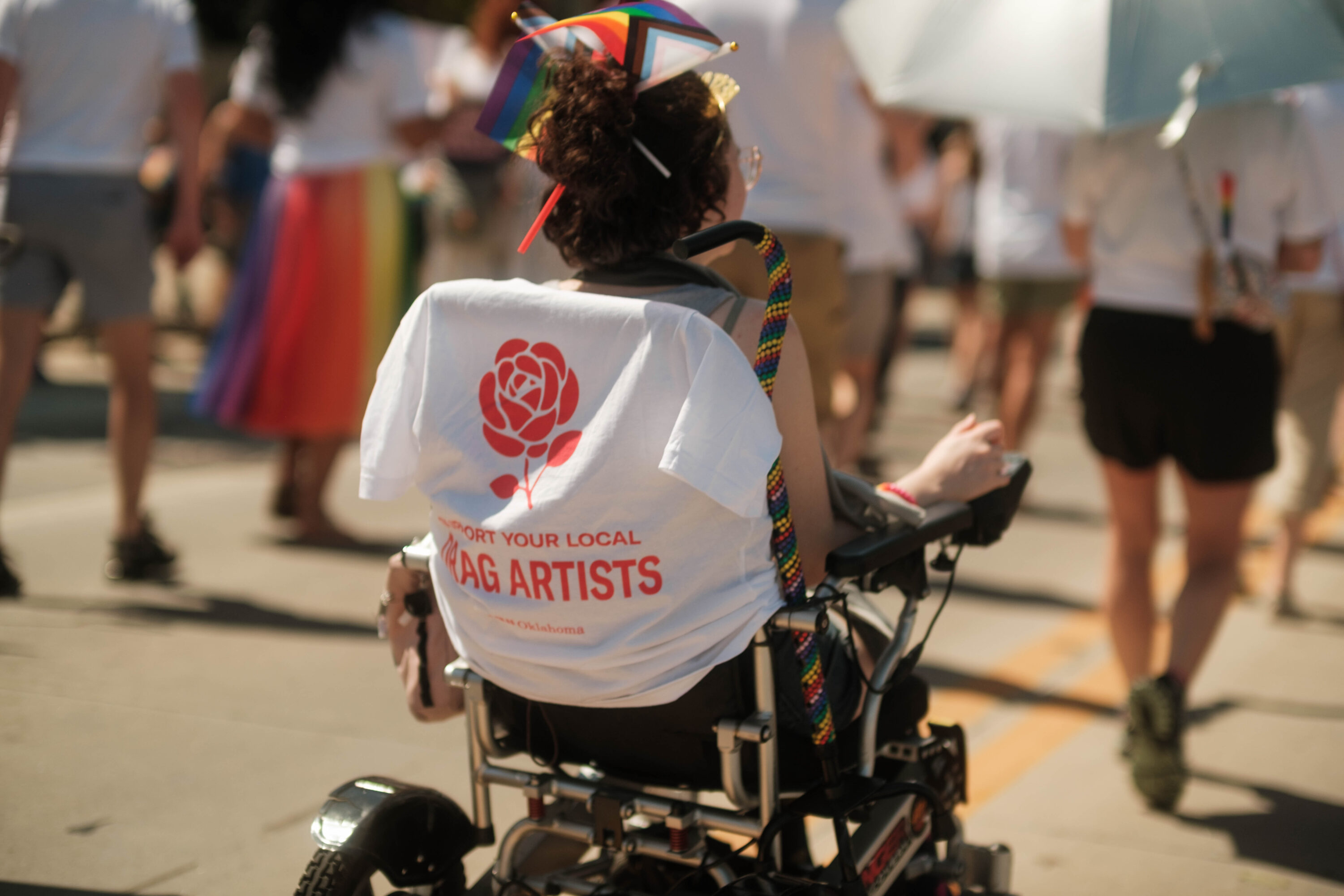 Photo of a person with pride flags in their hair and an ACLU tee-shirt that reads "Support your Local Drag Artist" wrapped around their wheelchair