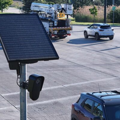 A solar-powered Flock camera is mounted on a pole beside a busy multi-lane road, with cars and a utility truck driving through the intersection in the background.