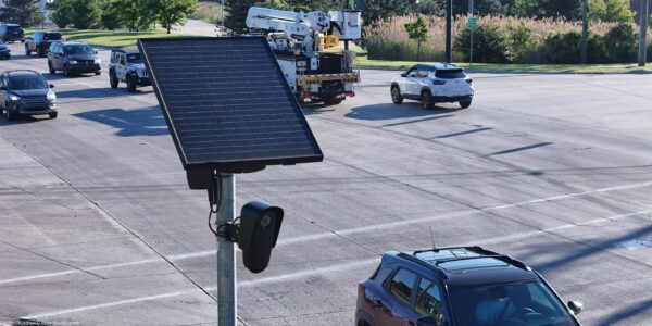 A solar-powered Flock camera is mounted on a pole beside a busy multi-lane road, with cars and a utility truck driving through the intersection in the background.