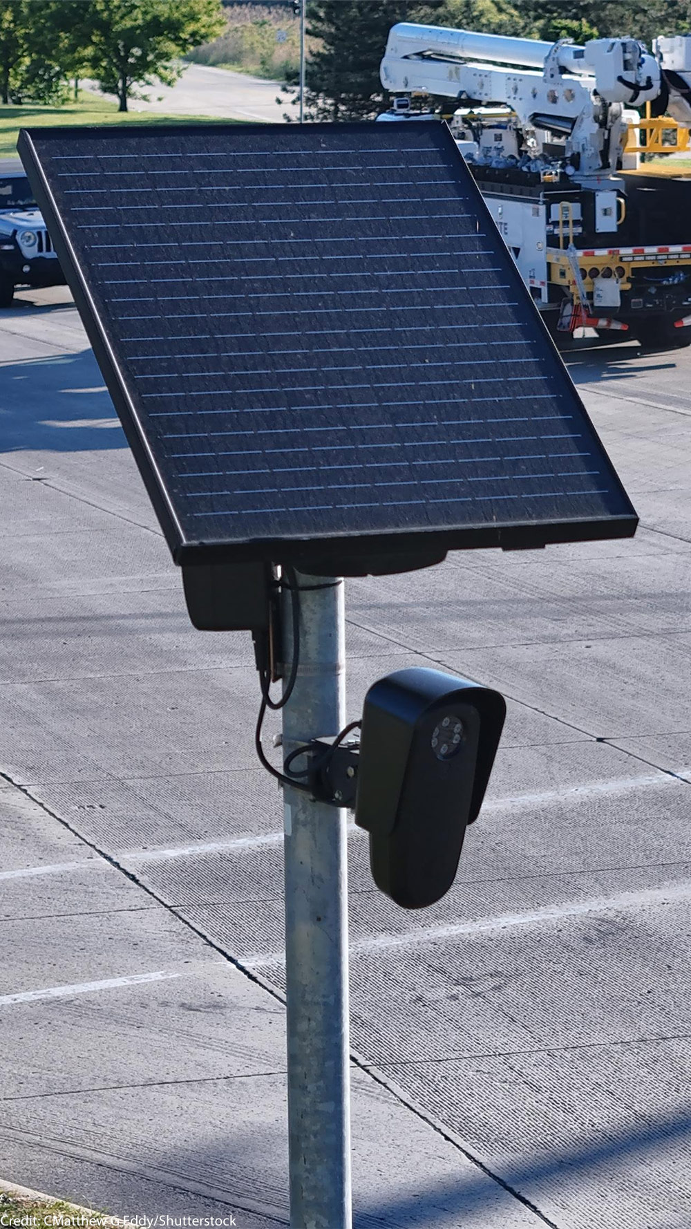 A solar-powered Flock camera is mounted on a pole beside a busy multi-lane road, with cars and a utility truck driving through the intersection in the background.