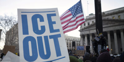 A hand holds a large white protest sign reading "ICE OUT" in bold blue letters during a crowded demonstration outside a government building. An American flag waves above the crowd, and additional signs in the background include messages calling to abolish ICE.