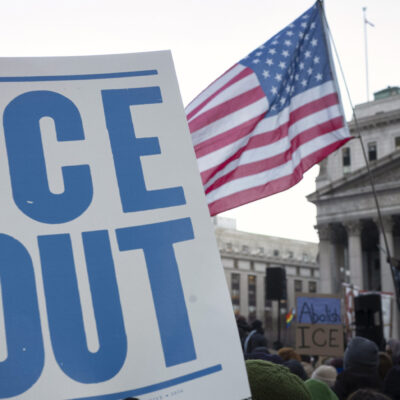 A hand holds a large white protest sign reading "ICE OUT" in bold blue letters during a crowded demonstration outside a government building. An American flag waves above the crowd, and additional signs in the background include messages calling to abolish ICE.