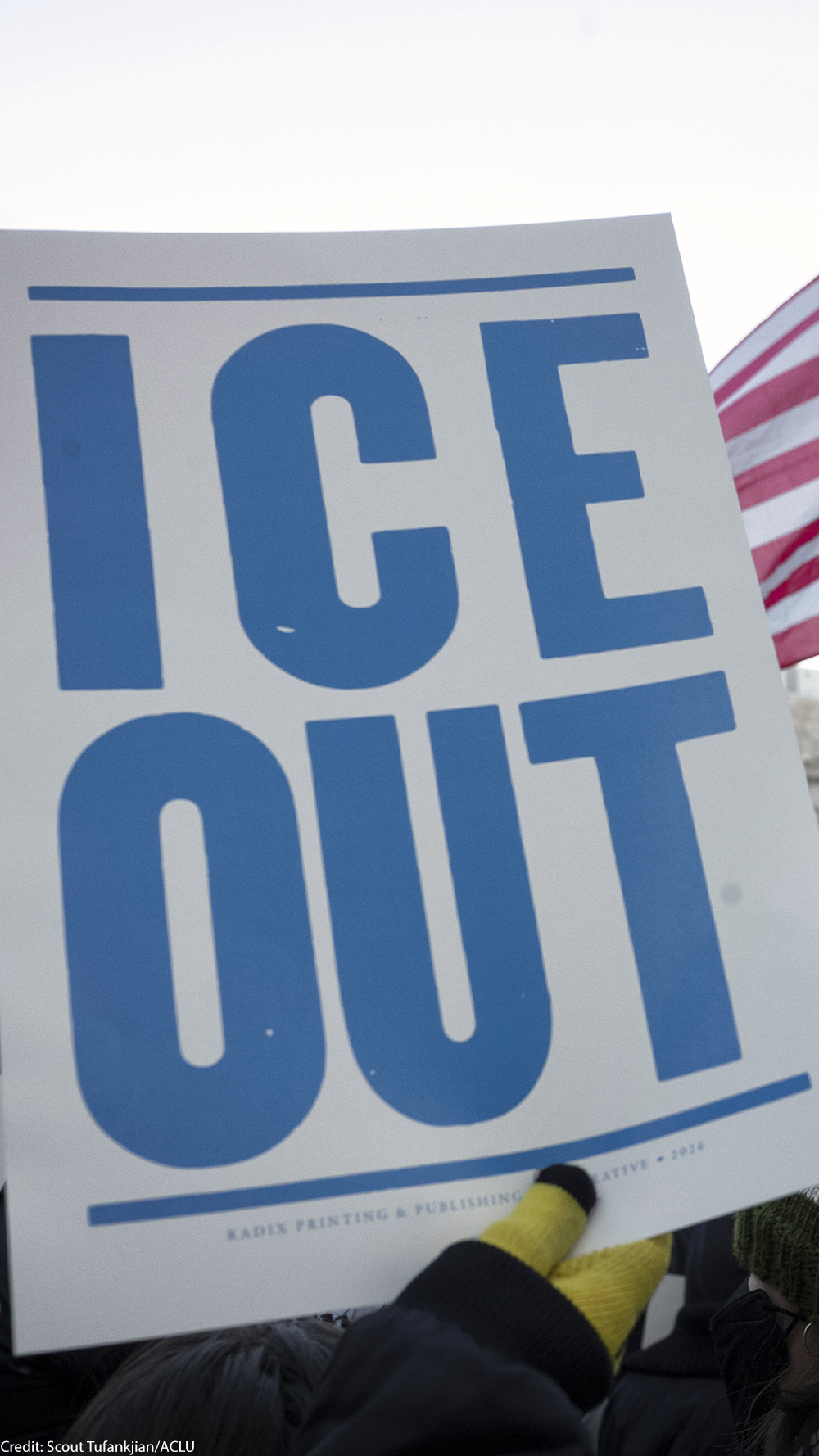 A hand holds a large white protest sign reading "ICE OUT" in bold blue letters during a crowded demonstration outside a government building. An American flag waves above the crowd, and additional signs in the background include messages calling to abolish ICE.
