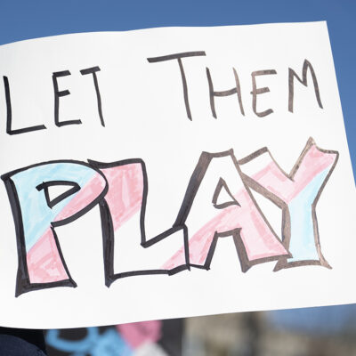 A hand holds a handmade sign reading "LET THEM PLAY," with the word "PLAY" colored in pink, blue, and white transgender pride colors, during a rally under a clear blue sky.