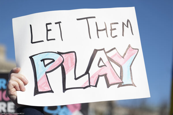 A hand holds a handmade sign reading "LET THEM PLAY," with the word "PLAY" colored in pink, blue, and white transgender pride colors, during a rally under a clear blue sky.