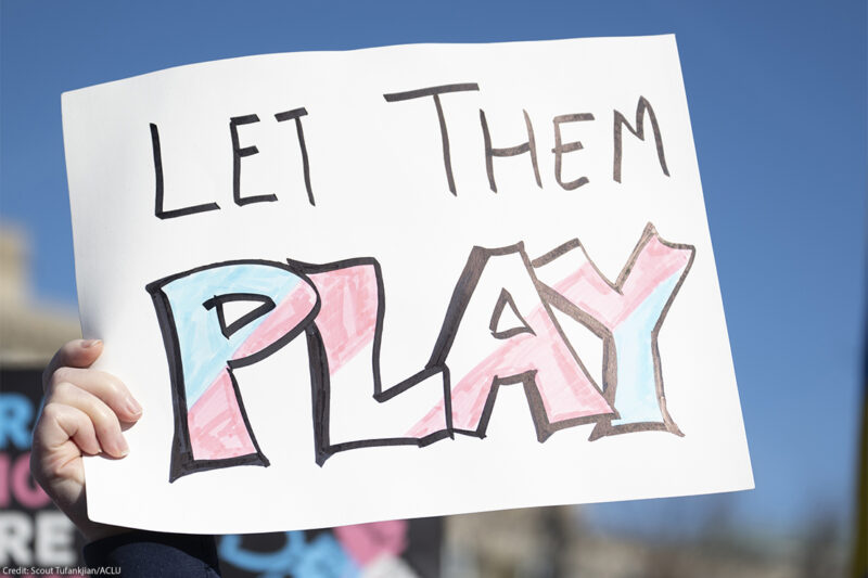 A hand holds a handmade sign reading "LET THEM PLAY," with the word "PLAY" colored in pink, blue, and white transgender pride colors, during a rally under a clear blue sky.