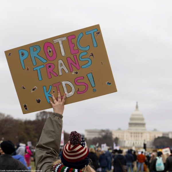 A demonstrator at a rally holds a handmade sign reading "Protect Trans Kids!" in pink, blue, and white letters, with the U.S. Capitol visible in the distance behind a large crowd on the National Mall.