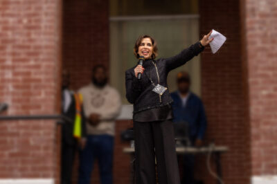 Yasmin Cader stands on an outdoor stage in front of a brick building, speaking into a microphone and gesturing with papers in her raised hand, while several people stand blurred in the background.