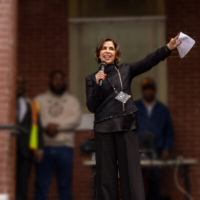Yasmin Cader stands on an outdoor stage in front of a brick building, speaking into a microphone and gesturing with papers in her raised hand, while several people stand blurred in the background.