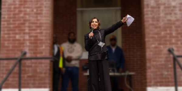 Yasmin Cader stands on an outdoor stage in front of a brick building, speaking into a microphone and gesturing with papers in her raised hand, while several people stand blurred in the background.