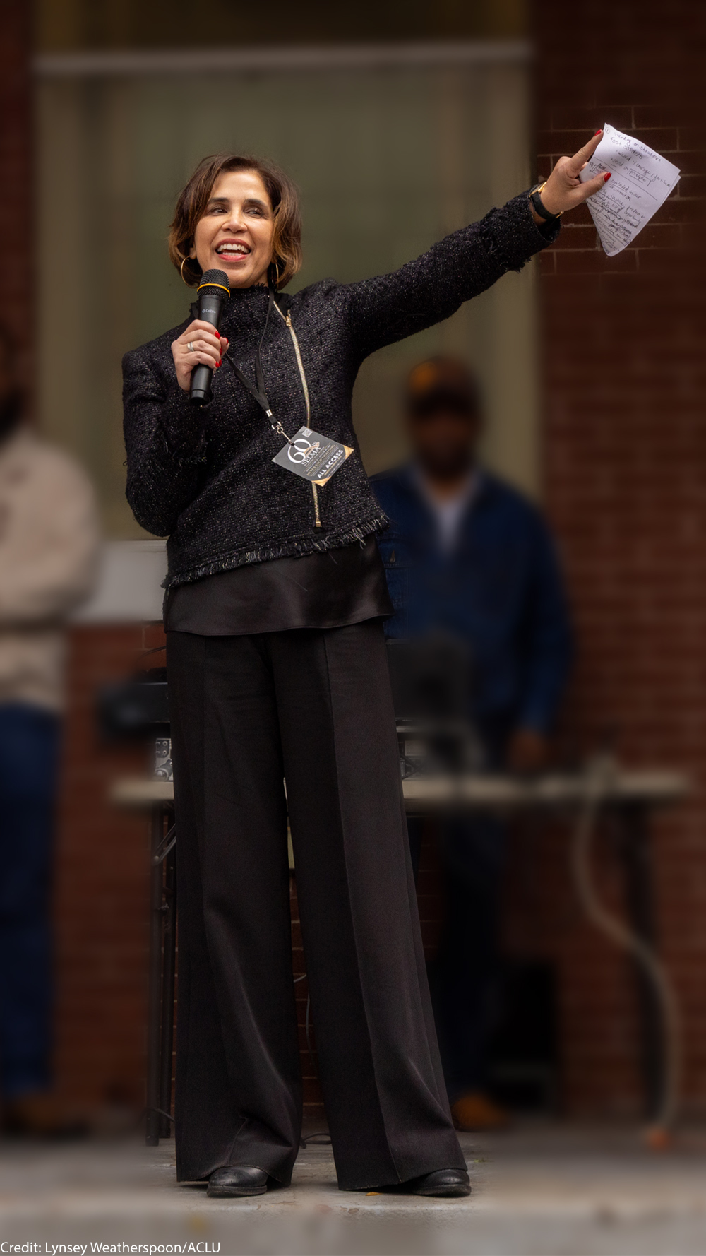 Yasmin Cader stands on an outdoor stage in front of a brick building, speaking into a microphone and gesturing with papers in her raised hand, while several people stand blurred in the background.