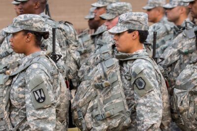 U.S. Army soldiers, including women in the foreground, stand in formation outdoors wearing digital camouflage uniforms, patrol caps, and large tactical backpacks with straps and attached gear, all facing the same direction.
