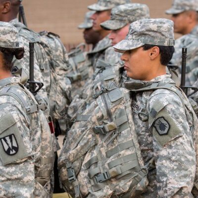 U.S. Army soldiers, including women in the foreground, stand in formation outdoors wearing digital camouflage uniforms, patrol caps, and large tactical backpacks with straps and attached gear, all facing the same direction.