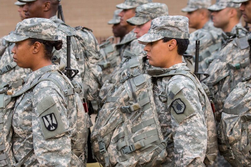 U.S. Army soldiers, including women in the foreground, stand in formation outdoors wearing digital camouflage uniforms, patrol caps, and large tactical backpacks with straps and attached gear, all facing the same direction.