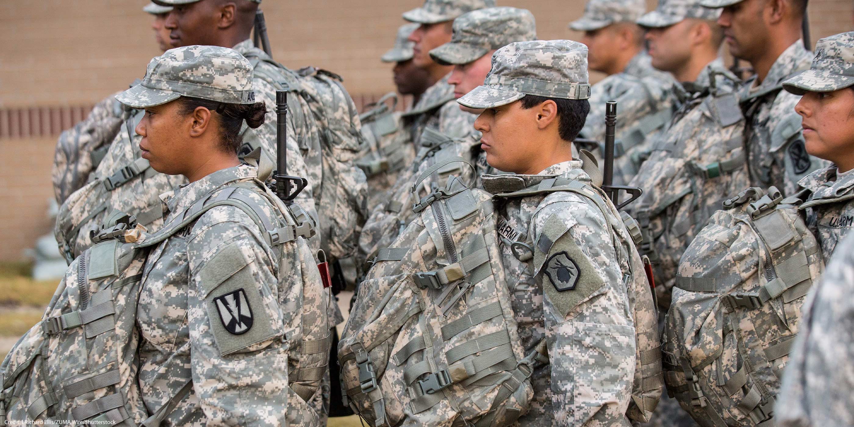 U.S. Army soldiers, including women in the foreground, stand in formation outdoors wearing digital camouflage uniforms, patrol caps, and large tactical backpacks with straps and attached gear, all facing the same direction.