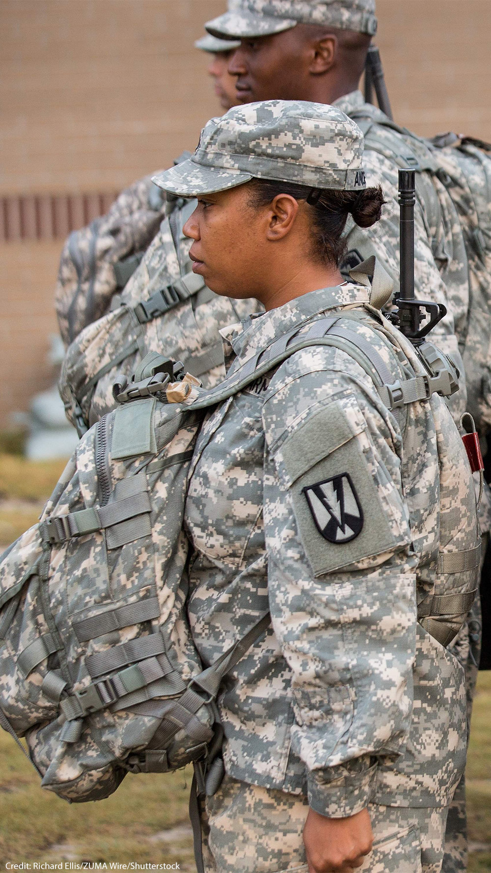 U.S. Army soldiers, including women in the foreground, stand in formation outdoors wearing digital camouflage uniforms, patrol caps, and large tactical backpacks with straps and attached gear, all facing the same direction.