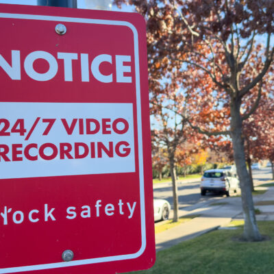 A red Flock Safety sign on a pole reads "Notice: 24/7 Video Recording" on a residential street, with parked cars, homes, trees, and a clear blue sky in the background.