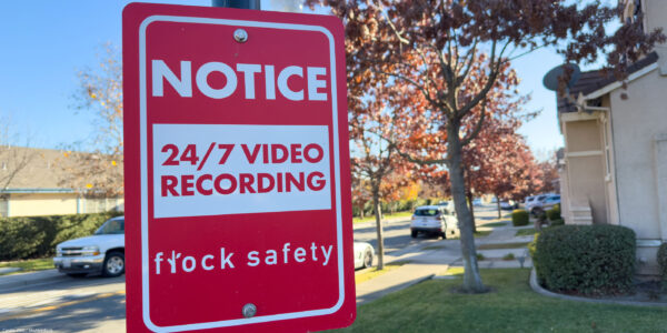 A red Flock Safety sign on a pole reads "Notice: 24/7 Video Recording" on a residential street, with parked cars, homes, trees, and a clear blue sky in the background.