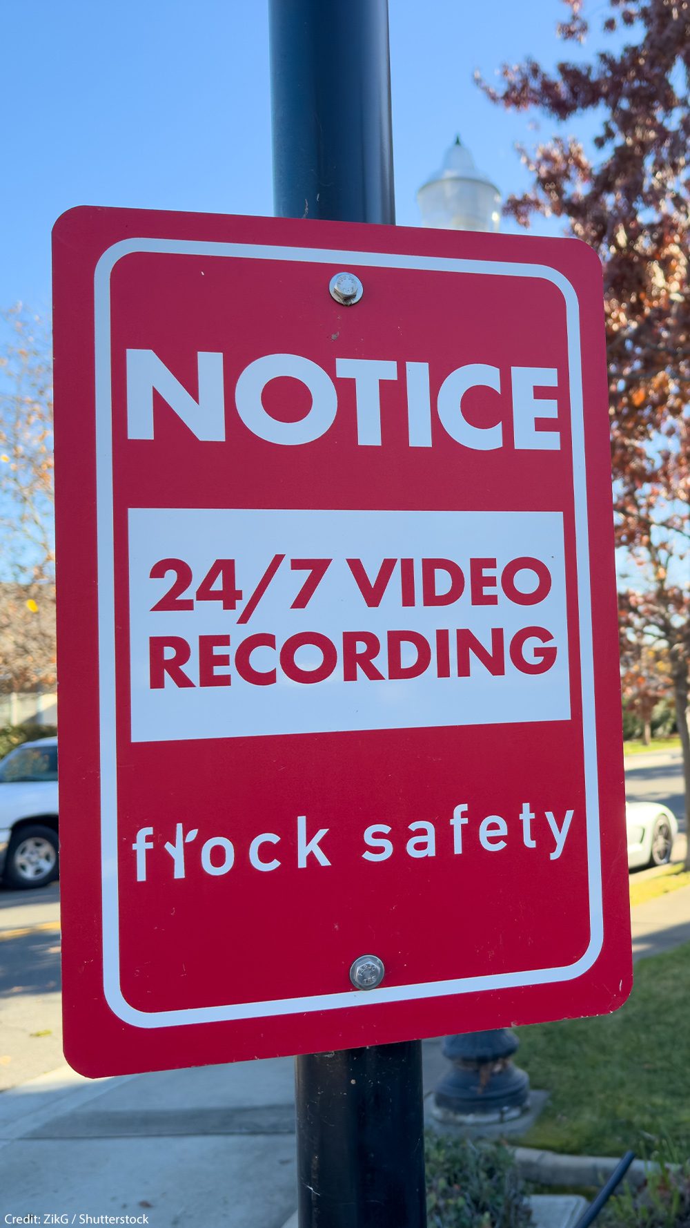 A red Flock Safety sign on a pole reads "Notice: 24/7 Video Recording" on a residential street, with parked cars, homes, trees, and a clear blue sky in the background.