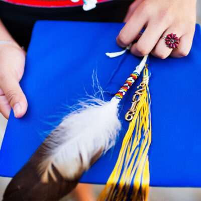 Close up image of a hand holding a graduation cap and tassel with traditional Navajo accessories and jewelry such as a beaded feather and coral ring.