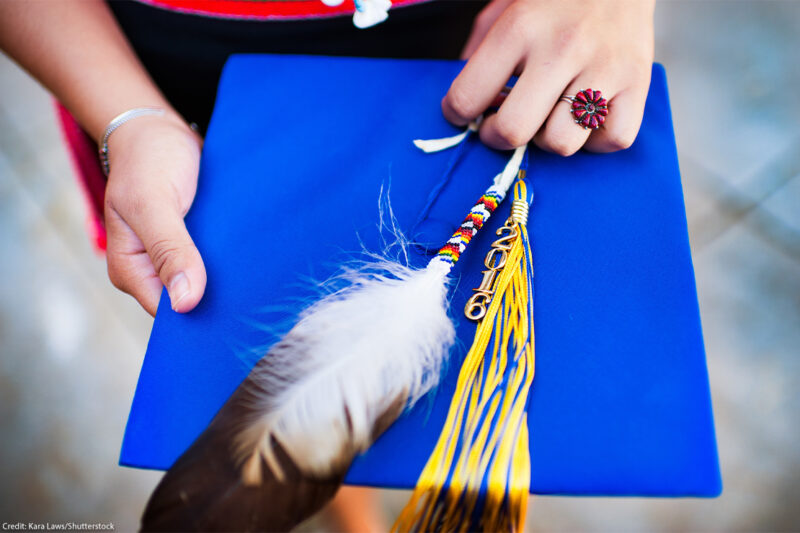 Close up image of a hand holding a graduation cap and tassel with traditional Navajo accessories and jewelry such as a beaded feather and coral ring.