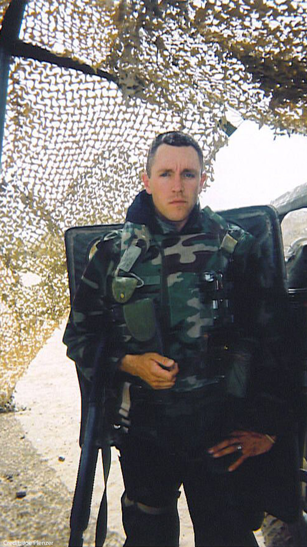 Joe Plenzer wearing a camouflage tactical vest stands beside a vehicle under camouflage netting in a sandy outdoor area.
