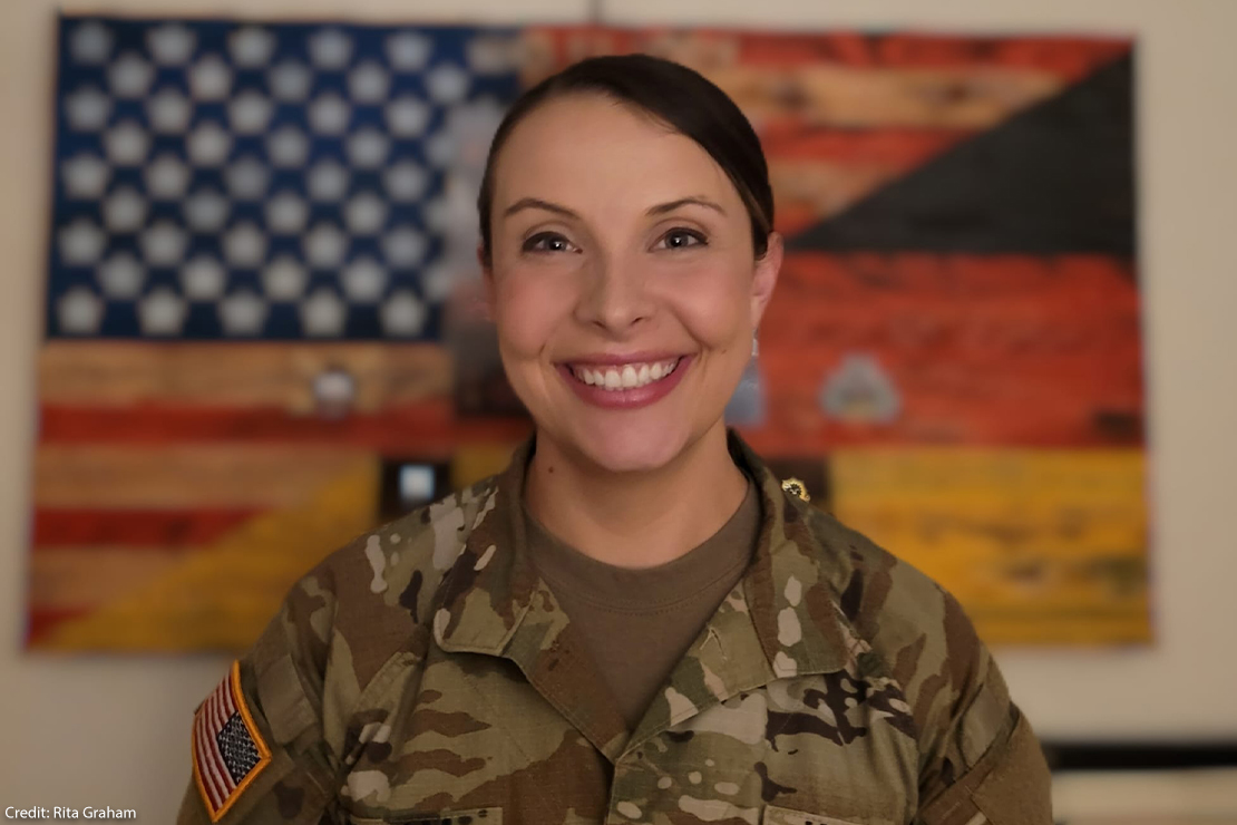 A smiling Rita Graham in a military camouflage uniform stands for a portrait, with an American flag artwork blurred in the background.