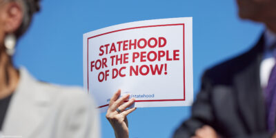 A person holds a white sign with bold red text and a red border reading "Statehood for the People of DC Now!" against a bright blue sky, with two blurred people in the foreground.