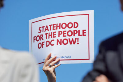 A person holds a white sign with bold red text and a red border reading "Statehood for the People of DC Now!" against a bright blue sky, with two blurred people in the foreground.