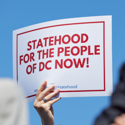 A person holds a white sign with bold red text and a red border reading "Statehood for the People of DC Now!" against a bright blue sky, with two blurred people in the foreground.
