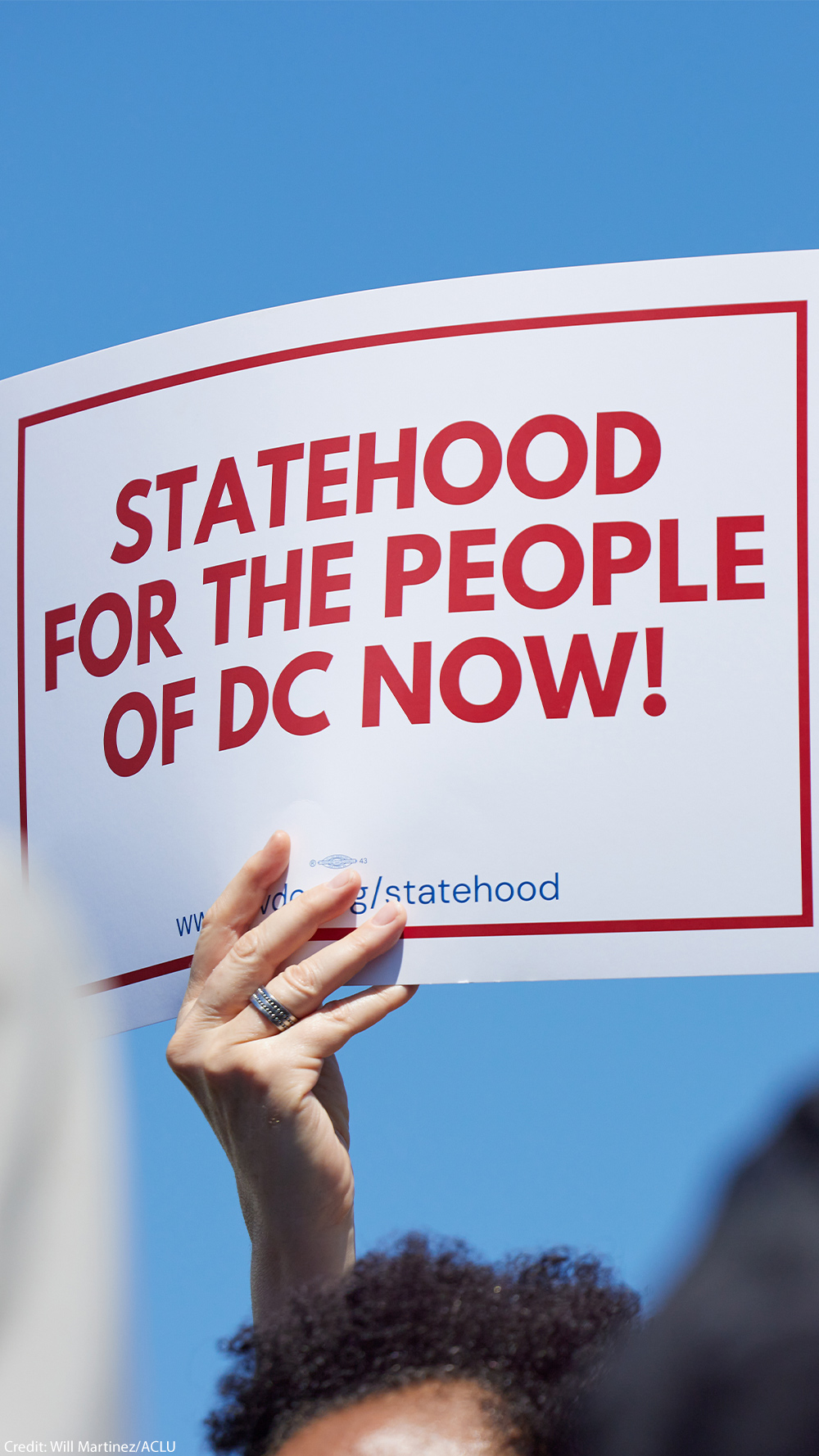 A person holds a white sign with bold red text and a red border reading "Statehood for the People of DC Now!" against a bright blue sky.