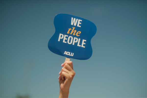 A hand, raised in the air, holding a small picket sign that reads "WE THE PEOPLE," with the ACLU logo.