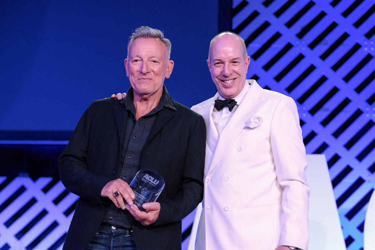 Bruce Springsteen stands onstage in a black suit next to ACLU Executive Director Anthony D. Romero, who smiles with his hand on Springsteen’s shoulder as he accepts the award. Romero is a light-skinned Puerto Rican man and wears a white dinner jacket and black bowtie.