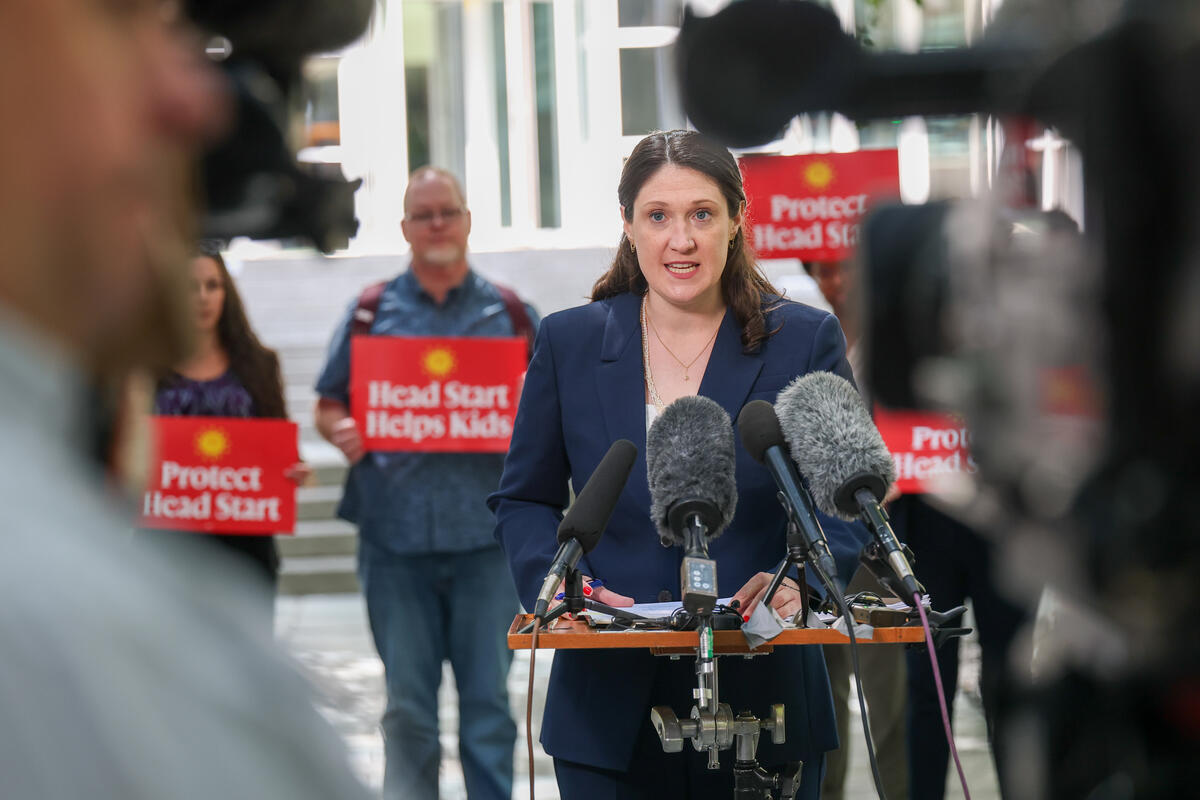 Jennesa Calvo-Friedman wears a navy suit and speaks into microphones before a press conference crowd
