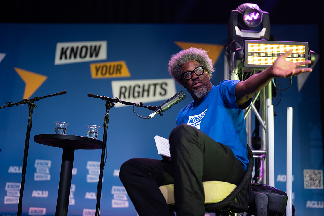 W. Kamau Bell on stage with a microphone at ACLU's Know Your Rights Bus Tour