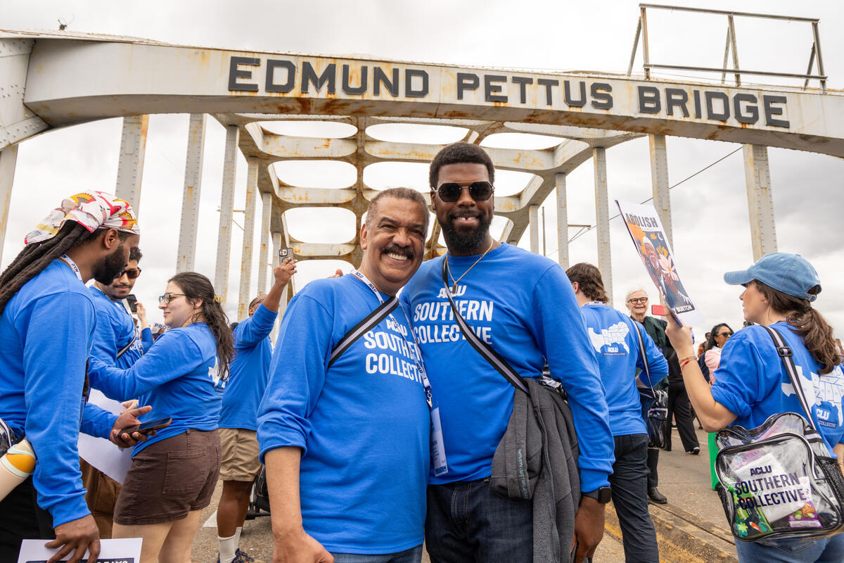 Two people wearing blue tee shirts that read "ACLU Southern Collective" pose on the Edmund Pettus Bridge, surrounded my other protestors